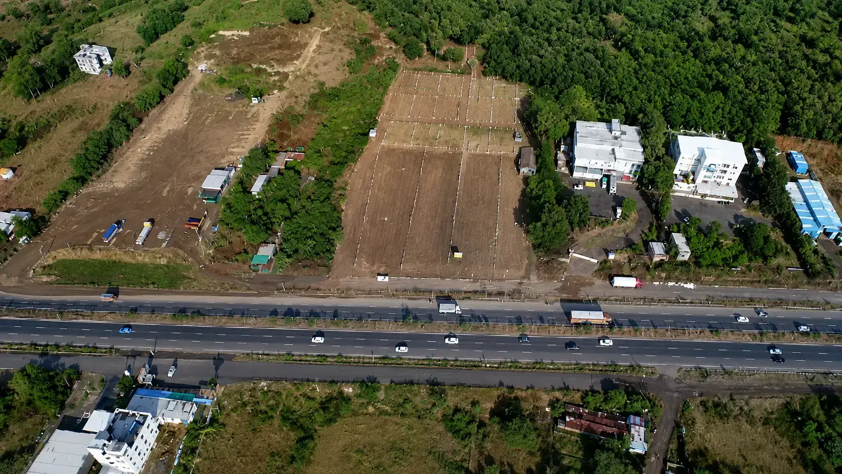 Aerial view of a major multi-lane highway running alongside cleared industrial plots and commercial buildings bordering a lush green landscape in the Khed Shivapur area.